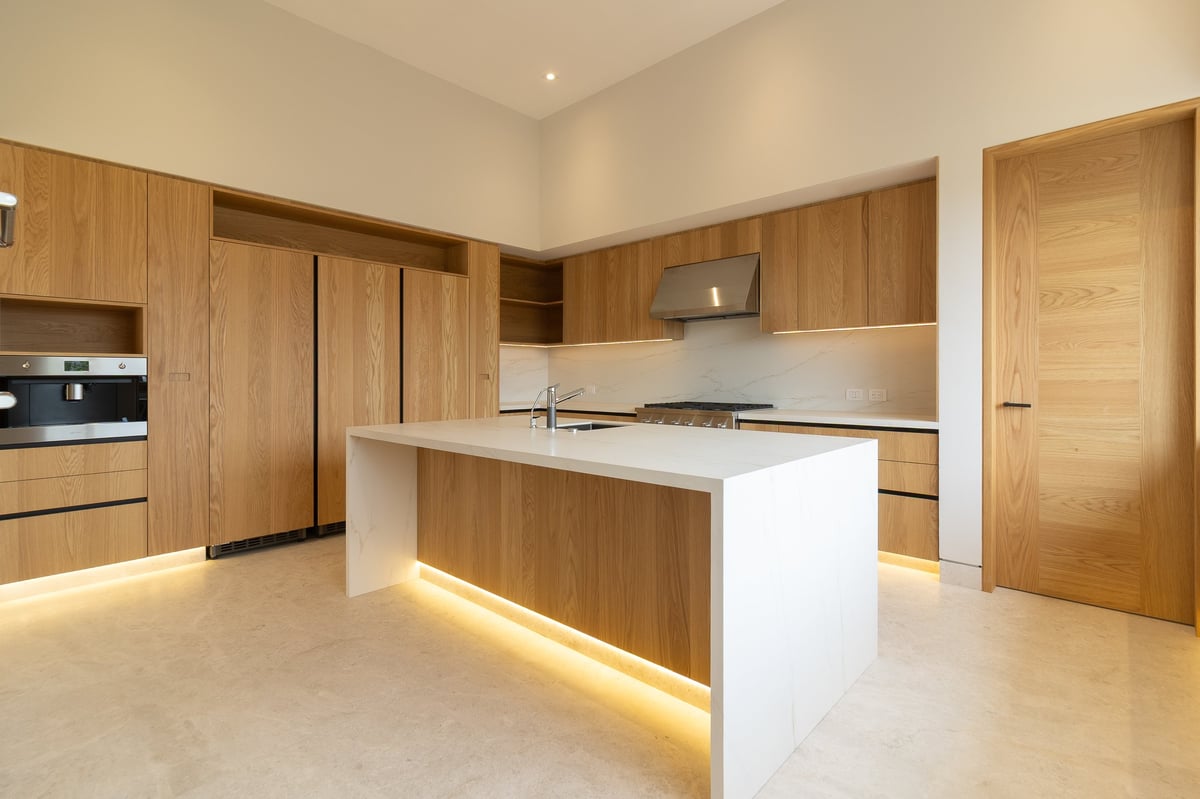 Modern kitchen with wood cabinetry, white waterfall island, warm ambient lighting, and light stone flooring