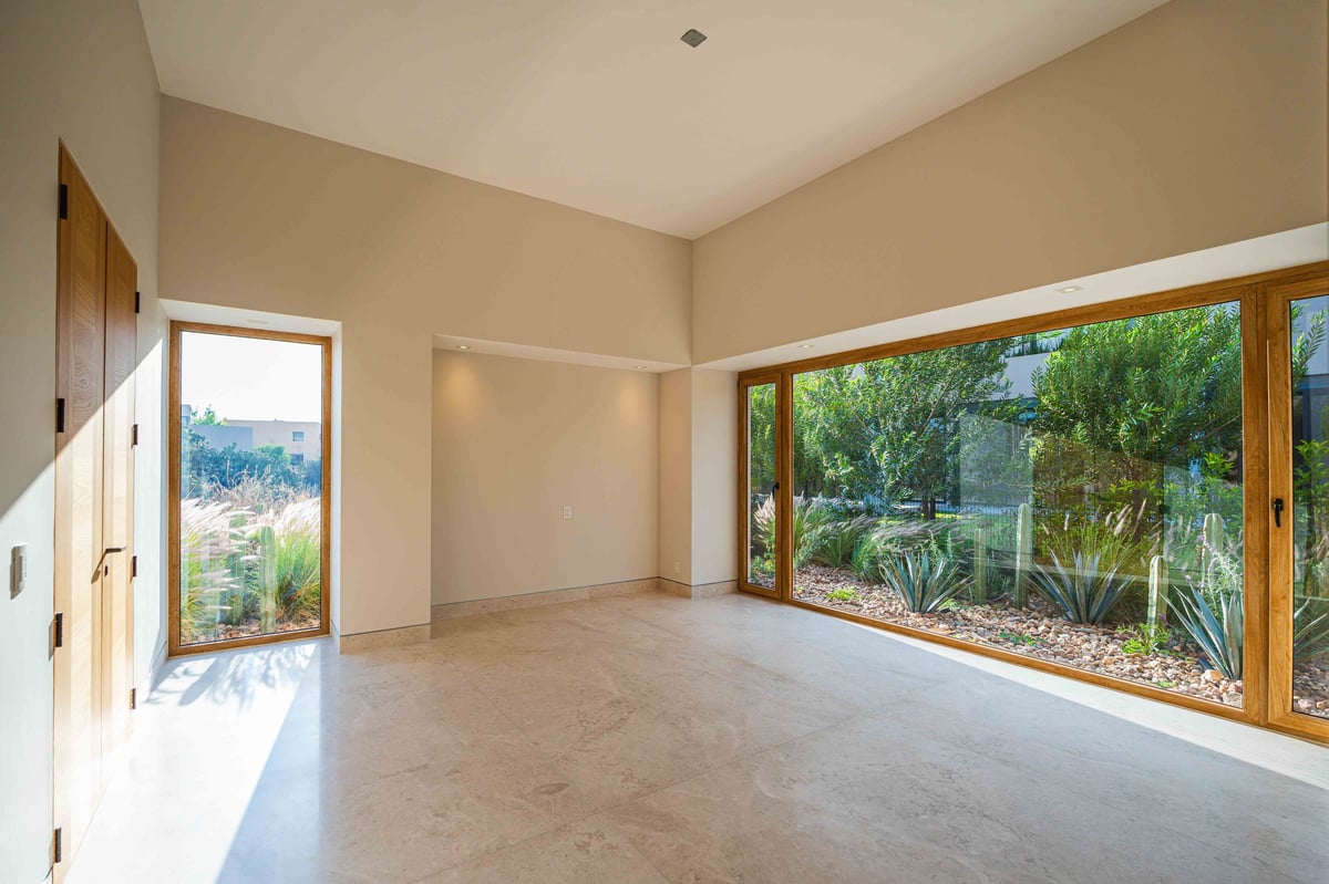 Empty modern room with beige walls, concrete floor, and large windows overlooking green landscape and trees