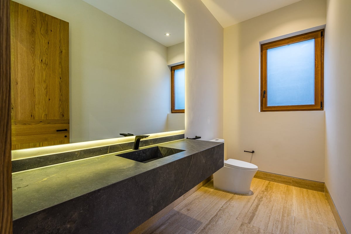 Modern bathroom with long black concrete sink, wooden cabinetry, toilet, and windows with wooden frames and warm wood flooring