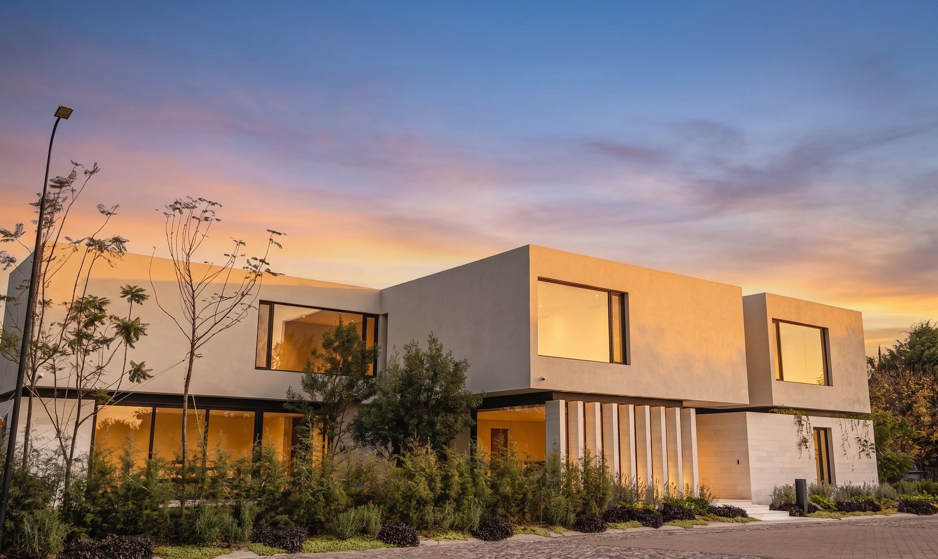 Modern minimalist white house with large windows at sunset, surrounded by landscaping and a gravel driveway