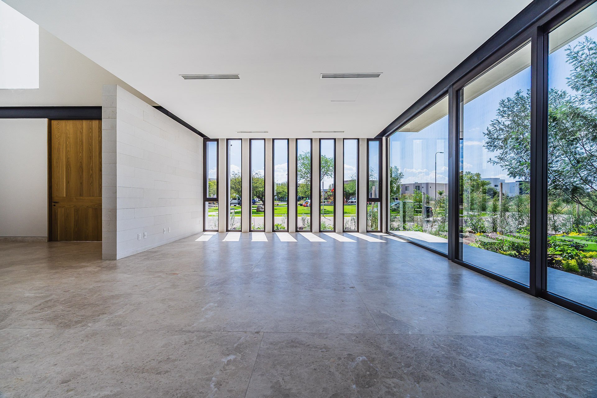 Modern empty room with polished concrete floors, white walls, wooden door, and large glass doors opening to outdoor garden views.
