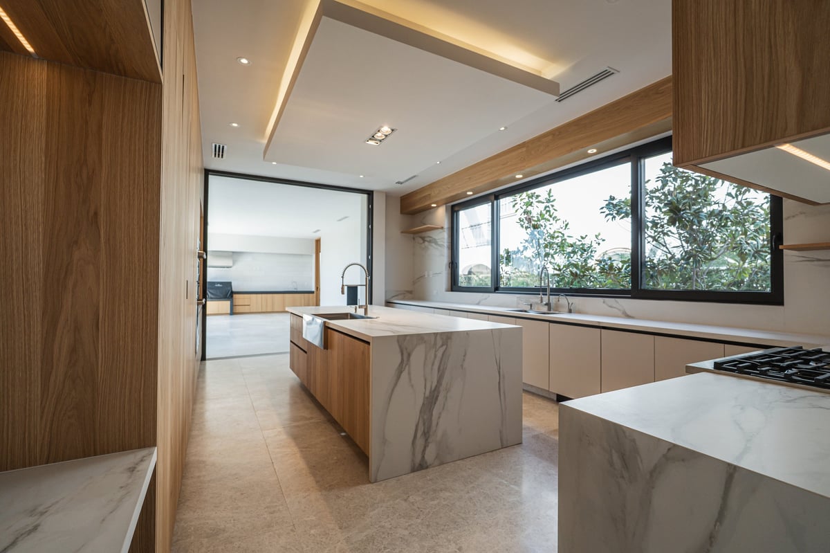 Modern kitchen with wooden cabinetry, marble island countertop, stainless steel appliances, and large windows overlooking greenery