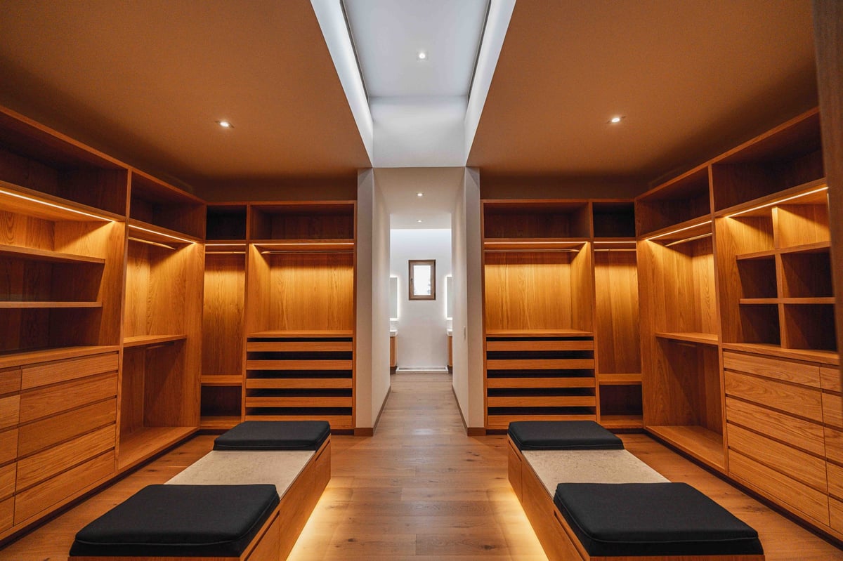 Luxury walk-in closet with wooden cabinetry, built-in shelving, and seating areas on both sides of a central aisle
