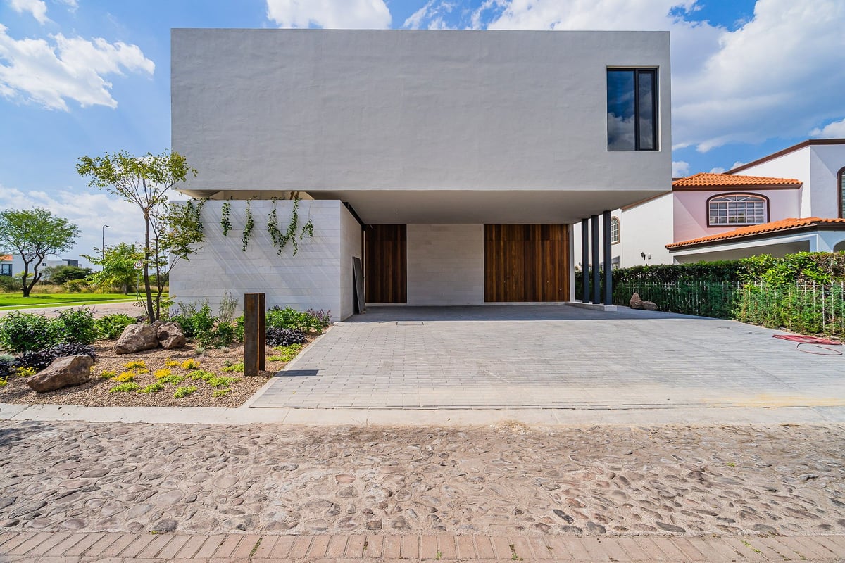 Modern two-story white house with minimalist design, wooden entrance, driveway, and landscaped front yard under blue sky