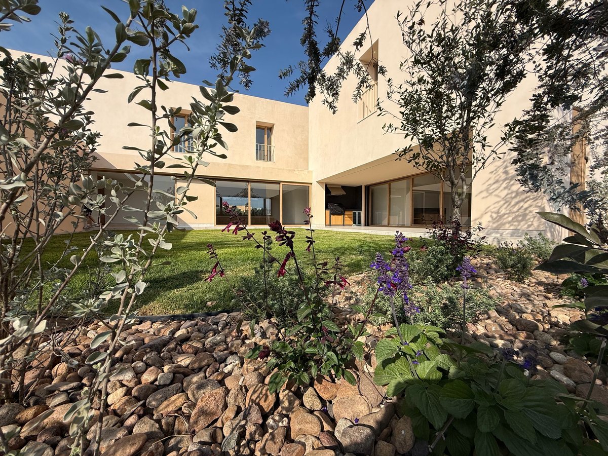 Modern beige house with landscaped garden featuring purple flowers, stone pathways, and mature trees on a sunny day
