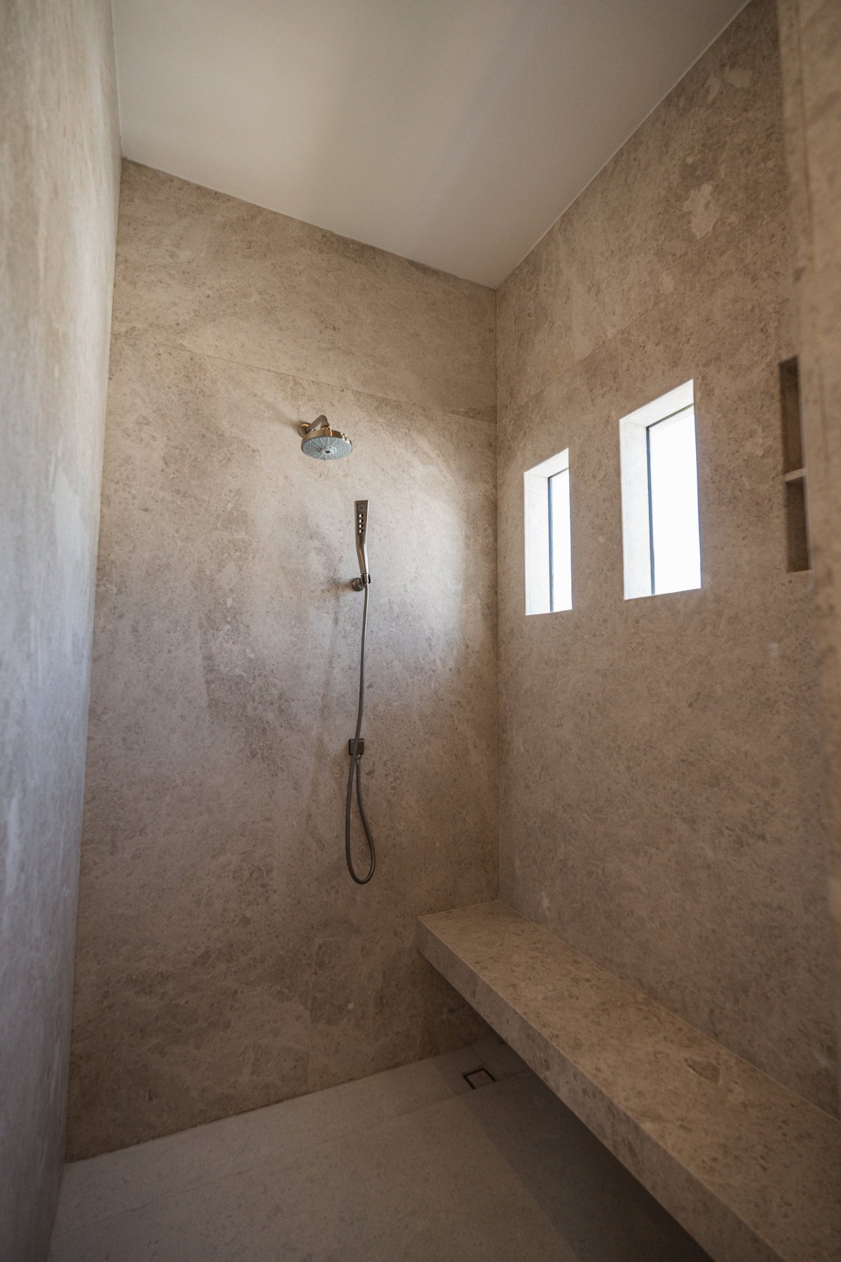 Modern bathroom shower enclosure with travertine tile walls, rain showerhead, handheld spray, built-in bench, and two rectangular windows