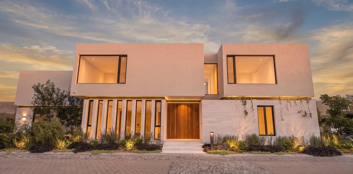 Modern white house with flat roof, geometric design, illuminated entrance, and landscaping at dusk