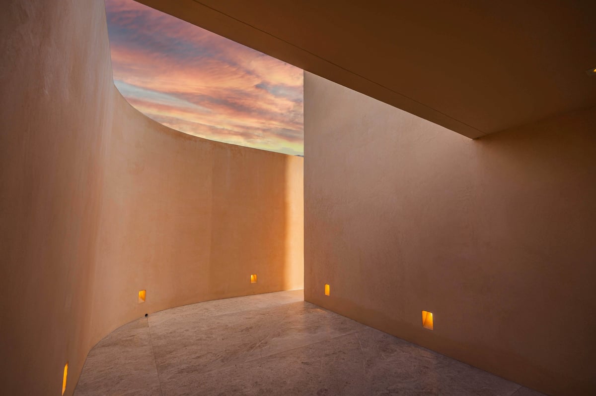 Modern minimalist interior with curved sand floor, warm beige walls, sunset sky window, and small glowing lights along the floor