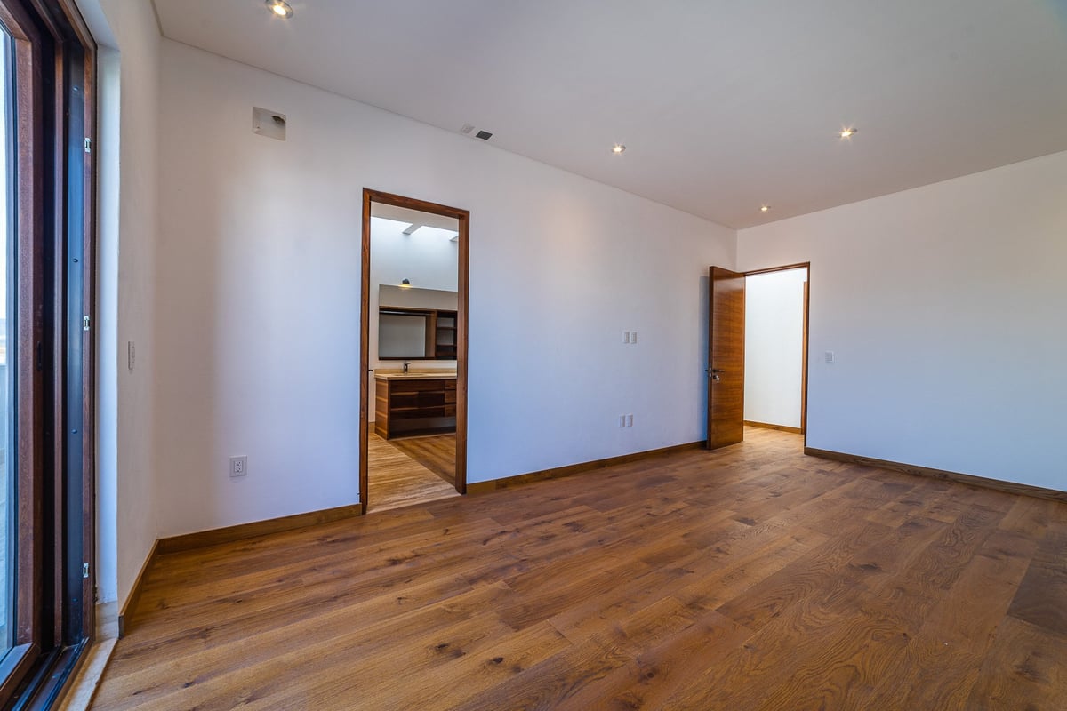 Empty modern bedroom with wooden floors, white walls, recessed lighting, and doorways leading to hallway and closet