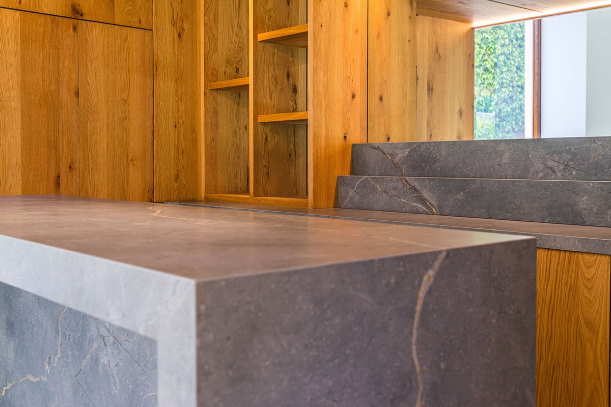 Modern kitchen with dark slate countertop and wooden cabinetry with shelving units and windows in background