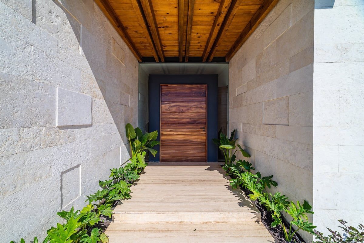 Modern entryway with wooden ceiling and horizontal slat door flanked by white stone walls and green plants along wooden floor