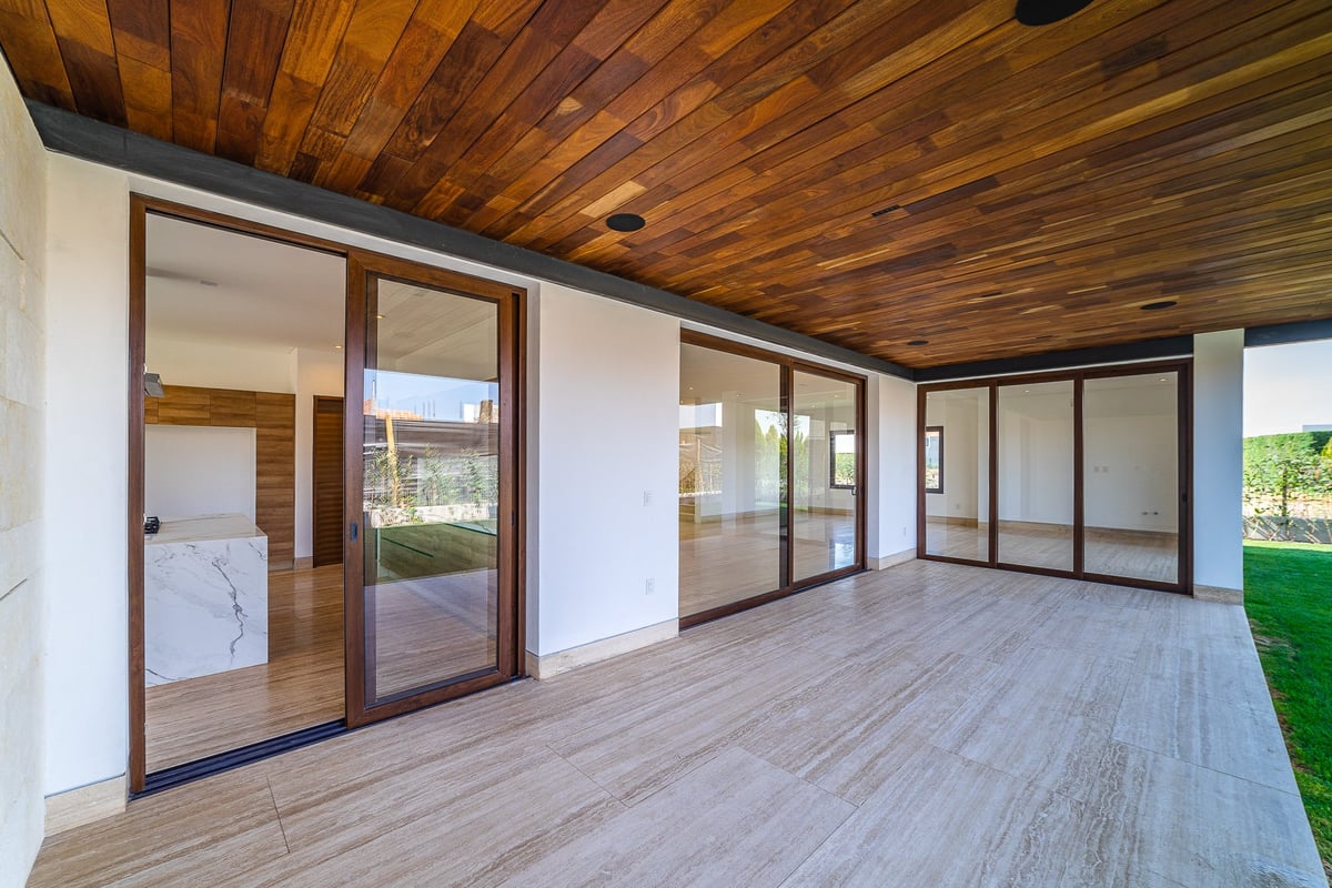 Empty modern living space with wooden ceiling, large glass sliding doors, light wood flooring, and white walls opening to outdoor views